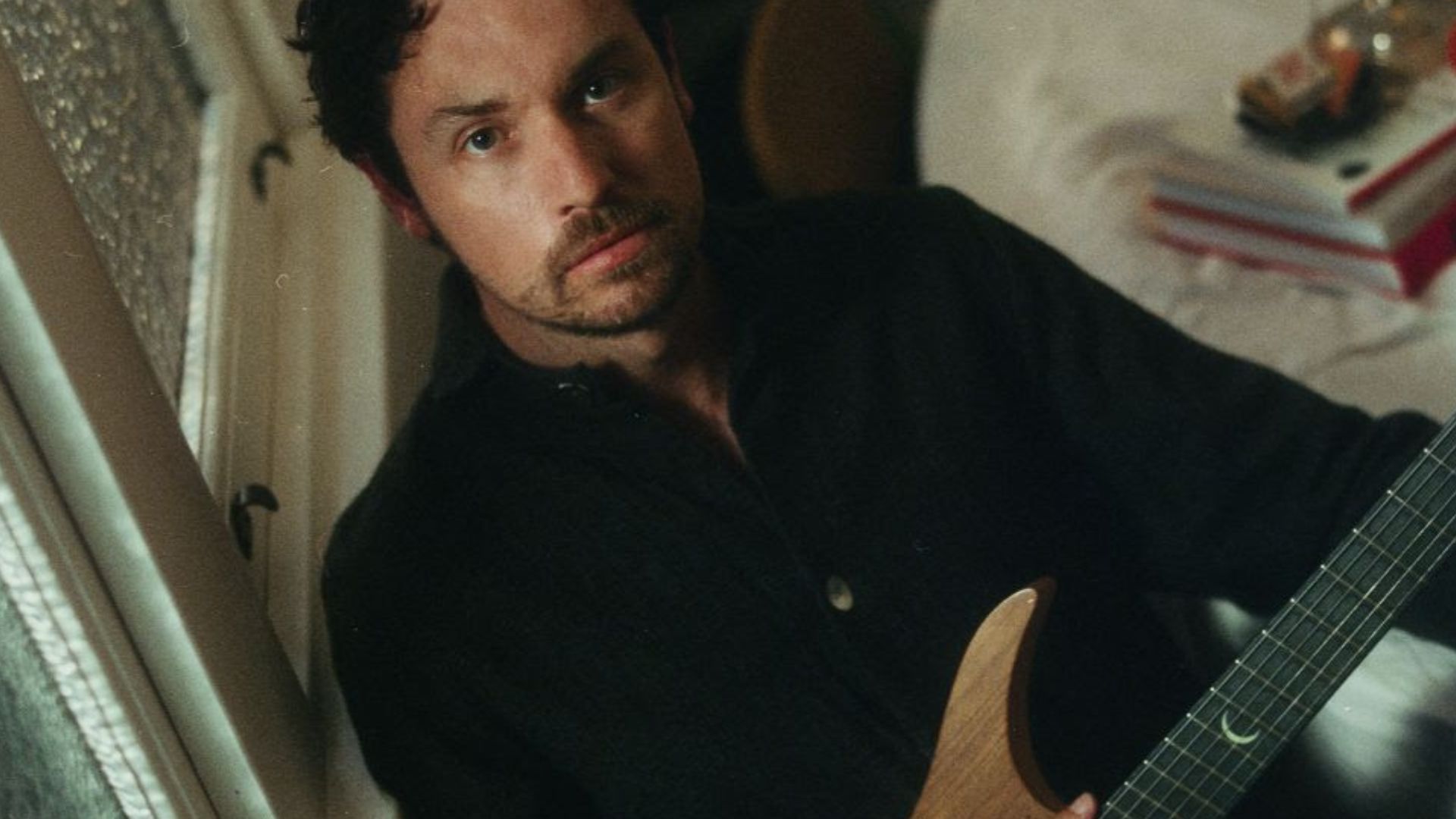 Plini reclines beside a window holding a guitar.