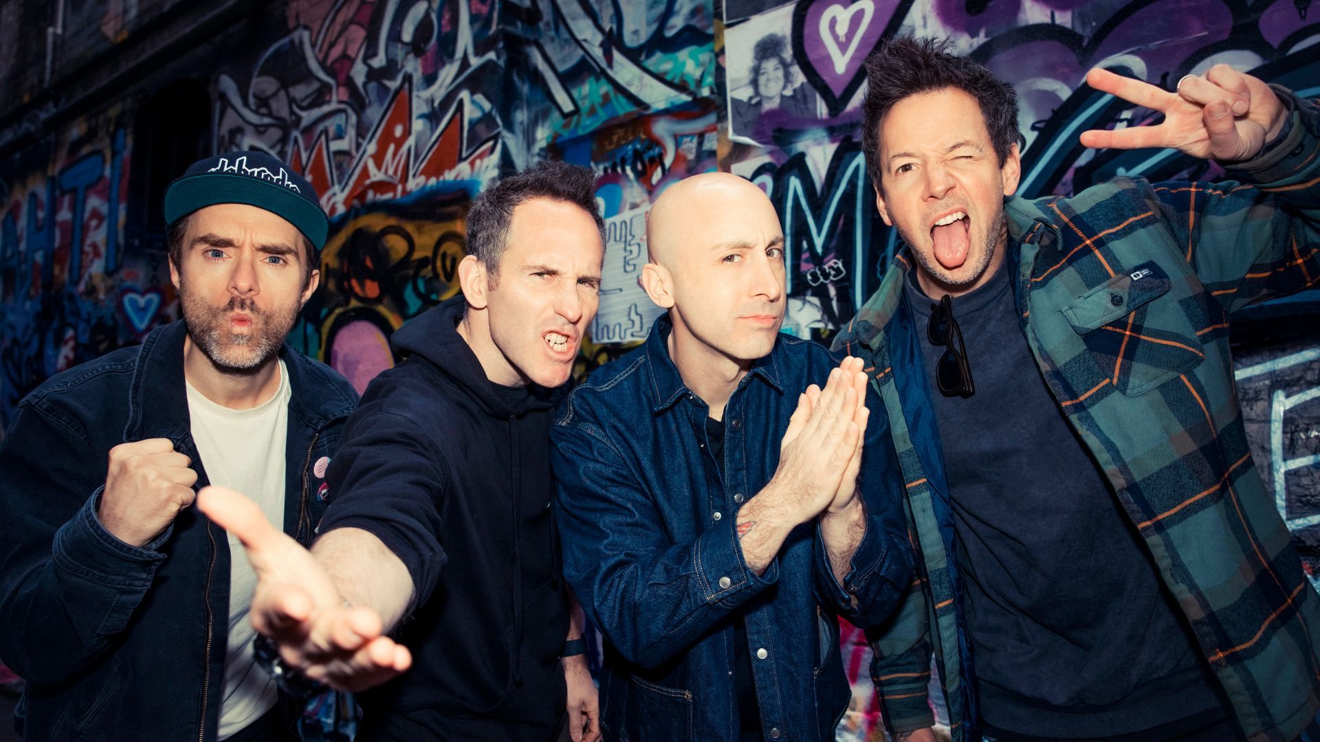 The four members of Simple Plan pose in front of a graffiti-covered wall.